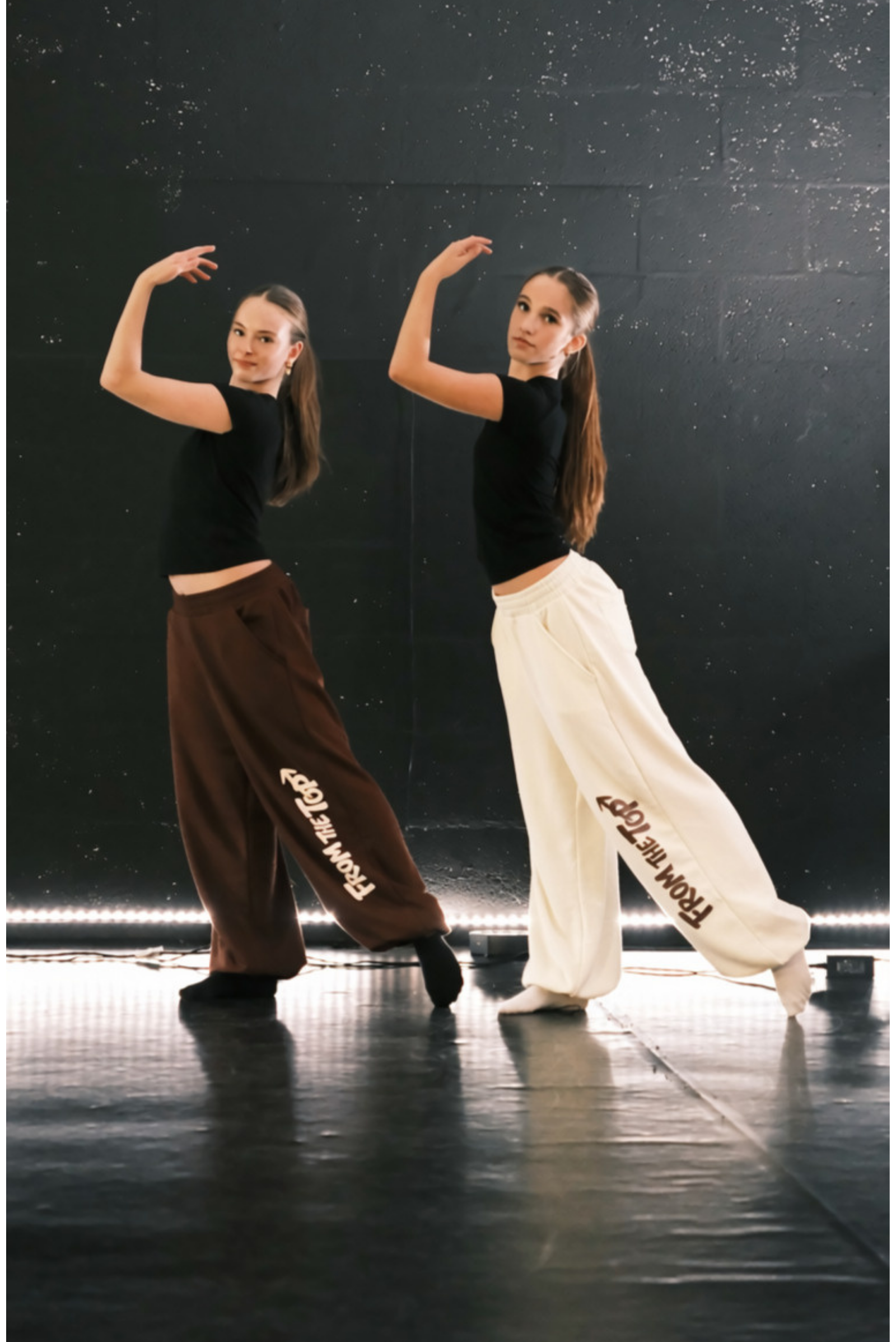 Two young dancers posing in a dance studio, wearing matching black crop tops and wide-leg sweatpants in brown and off-white, both featuring the text 'From the top'  printed down the side.