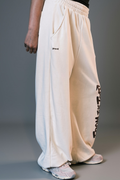 A person wearing loose off-white sweatpants with black graphic text down one leg and a small black logo on the other leg, paired with light-colored sneakers against a gray studio background.