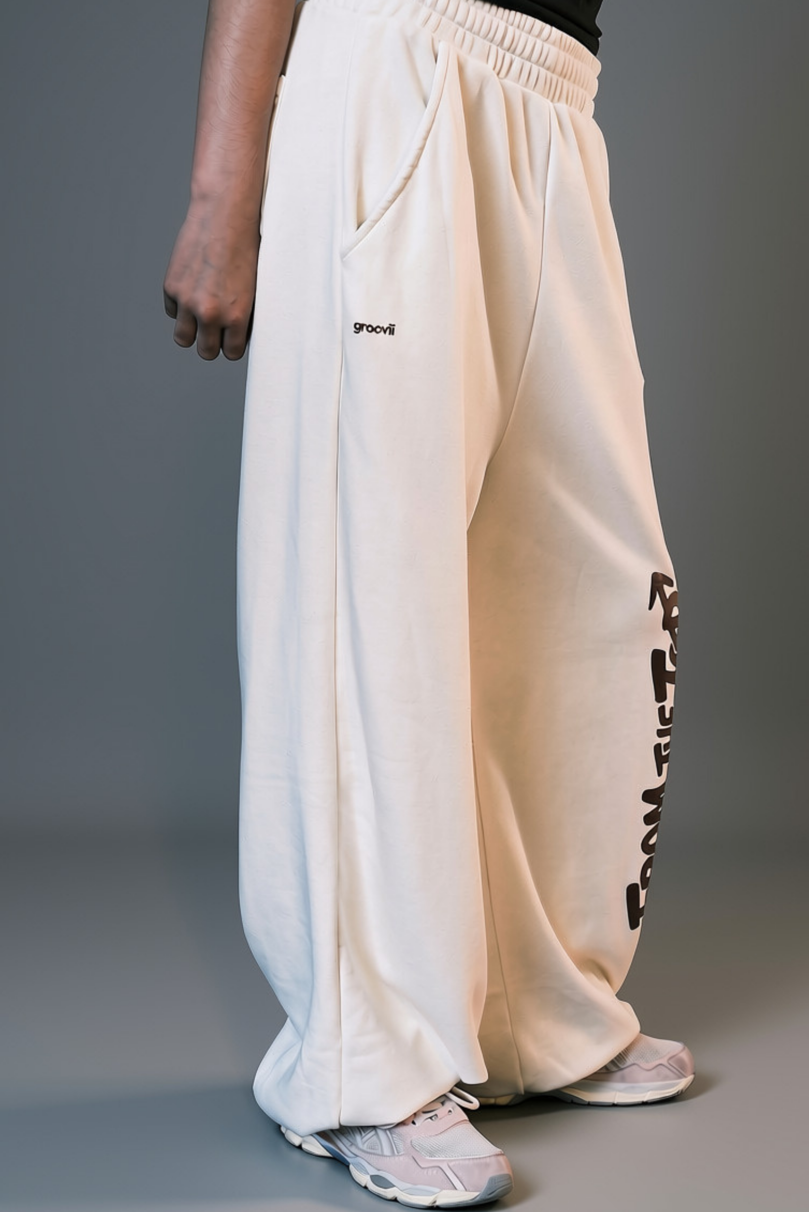 A person wearing loose off-white sweatpants with black graphic text down one leg and a small black logo on the other leg, paired with light-colored sneakers against a gray studio background.